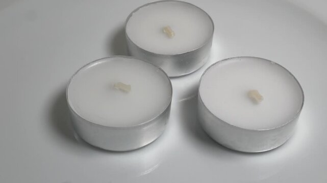 Close-up of three white tea light candles in silver aluminum cups on a white plate, captured on a rotating surface against a neutral grey background.