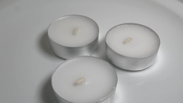 Close-up of three white tea light candles in silver aluminum cups on a white plate, captured on a rotating surface against a neutral grey background.