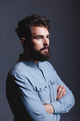 Obraz premium Studio portrait of a serious, confident young bearded man standing with arms crossed, looking in profile to the right against a solid dark grey background with copy space.