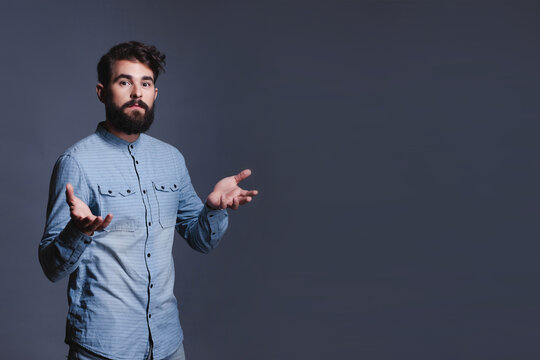 Studio portrait of a confused young bearded man shrugging with open arms and copy space.