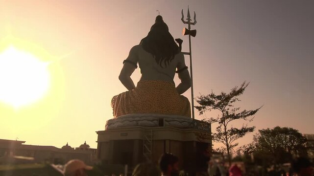 Silhouette of Lord Shiva statue with trishul near a temple during golden sunset in India. Spiritual Hindu scene with devotees, dramatic sky, Chaar Dham Temple in Vrindavan, Uttar Pradesh.