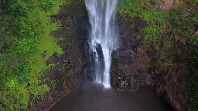 Aerial view shows a tall waterfall plunging into a calm pool. Surrounding cliffs are covered in thick, vibrant green moss and foliage. Water cascades powerfully down dark, wet rock faces
