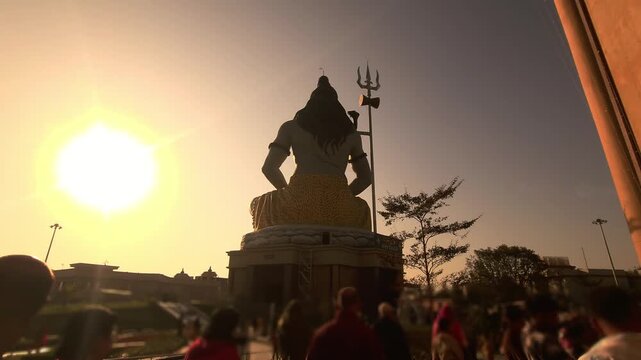 Silhouette of Lord Shiva statue with trishul near a temple during golden sunset in India. Spiritual Hindu scene with devotees, dramatic sky, Chaar Dham Temple in Vrindavan, Uttar Pradesh.