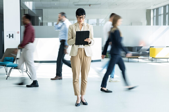 Businesswoman standing still and focusing on digital tablet, ignoring motion blurred colleagues walking fast around her in a modern office