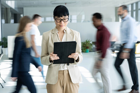 Businesswoman standing with a digital tablet, managing tasks while other employees are moving fast in a modern office environment