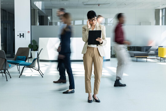 Professional businesswoman concentrating on her digital tablet in a busy, modern office lobby with people in motion blur
