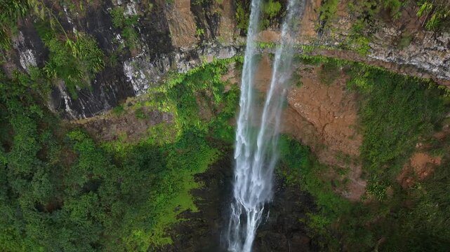 Aerial view of a majestic waterfall plunging down a steep, forested cliff. Water cascades in a powerful stream into a dark pool below. Lush green vegetation covers the cliffside, with mossy rocks