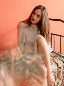 Stylish caucasian woman in boudoir dress sitting on bed with warm background and artistic light effects.