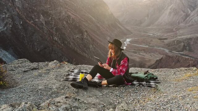 Young woman sitting on a rocky cliff using a tablet while overlooking a wide mountain valley with winding river below. Traveler combining outdoor adventure with digital lifestyle, working remotely or