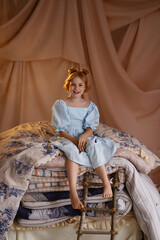Obraz premium Smiling Caucasian girl in blue dress sitting on stacked mattresses like princess and the pea, front view, look at camera, studio with beige drapery. Concept of fairy tale, childhood imagination.