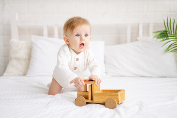 Fototapeta premium smiling baby 6 months old blond boy is sitting on a large bed in a bright bedroom and playing with a wooden toy car in a cotton bodysuit, the concept of children's goods