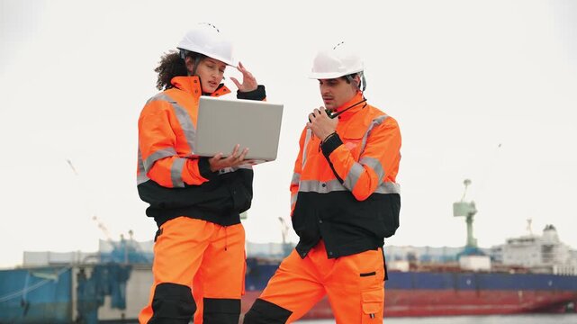 Logistics supervisors in safety uniforms using a laptop and walkie talkie at a commercial dock, managing maritime transport and supply chain operations near a large industrial cargo ship