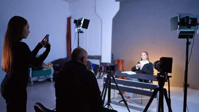 Cameraman and female assistant are filming the lady sitting at desk. Video footage in studio.