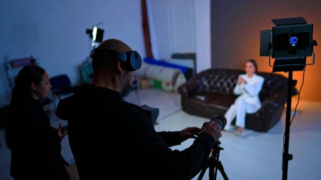 Man and woman work in the photo studio backstage. Lady wearing white suit sits on the sofa waiting for filming. Blurred backdrop.