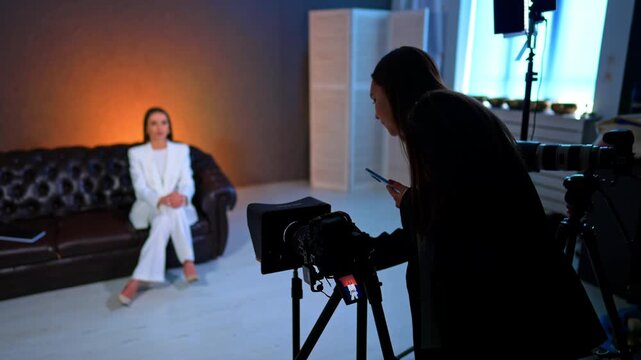 Woman with phone in hand checking the camera in tripod. Lady wearing white suit sits on the sofa in the backdrop. Photo studio backstage.