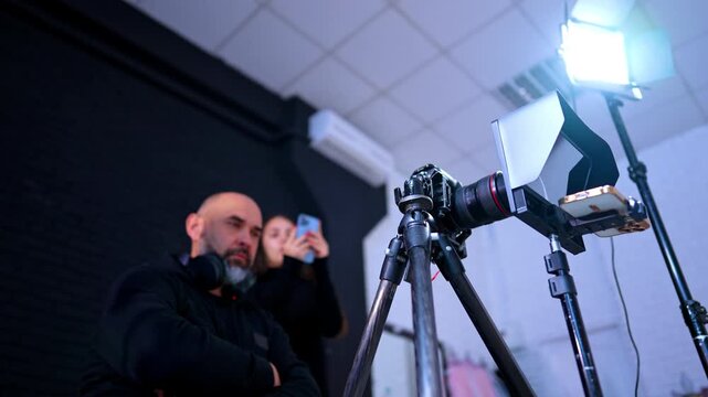Serious male cameraman in black hoodie sits in front of the camera. Low angle view. Woman holding a phone stands in the backdrop.
