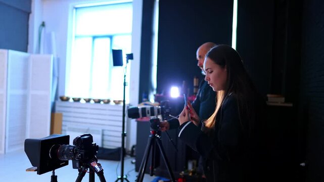 Long-haired woman uses phone to record video and man uses professional camera on tripod. Filming grew working backstage in photo studio.