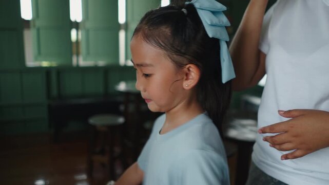An Asian girl smiles softly and shakes her head in front of a mirror while someone combs her hair