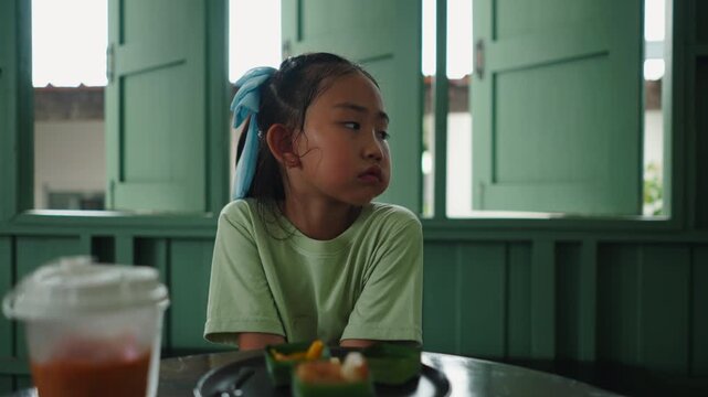 A little girl in a green shirt sits at a table to eat and drink with a blank stare near wooden windows