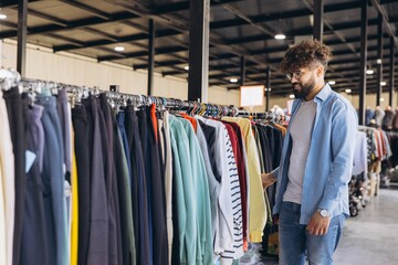 Obraz premium Arab man browsing clothing rail in a store, choosing new apparel, enjoying a shopping experience