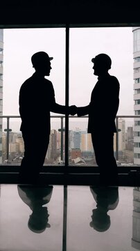 Silhouettes of two engineers in hard hats shaking hands inside a modern building with large windows and urban skyline in the background. Professionals confirming agreement, partnership or successful