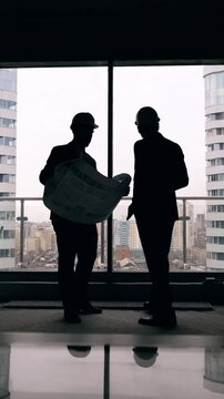 Silhouettes of two engineers in hard hats examining architectural blueprint inside a modern building with large windows and city view. Professionals discussing construction project, planning