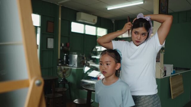 A little Asian girl dances in front of a mirror while her mother combs her hair with the help of a comb