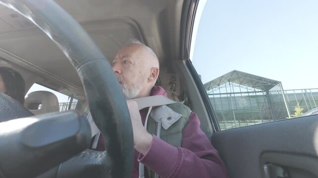 Low angle interior view of a middle aged bearded man driving a car. The driver is looking to the side while holding the steering wheel. Natural daylight enters through the window