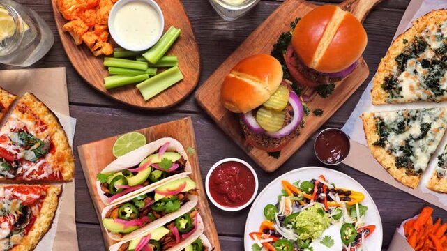 Healthy plant based fast food table scene. Slow rotating zoom motion over wood. Cauliflower crust pizzas, bean burgers, mushroom tacos, bell pepper nachos and cauliflower wings and sweet potato fries.