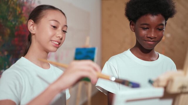 Diverse children in casual shirt painting canvas together in creative art lesson. Multicultural happy high school student drawing a same canvas while standing at colorful stained wall. Edification