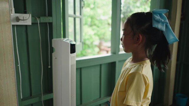 A young woman in a yellow shirt interacts with a white air cooler near a window by touching buttons