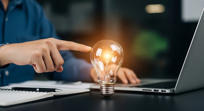 Person pointing at glowing light bulb on desk with laptop and notebook idea innovation inspiration creativity