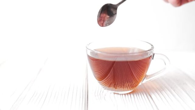 Close up of adding a teaspoon of white sugar into a hot drink in a glass mug.