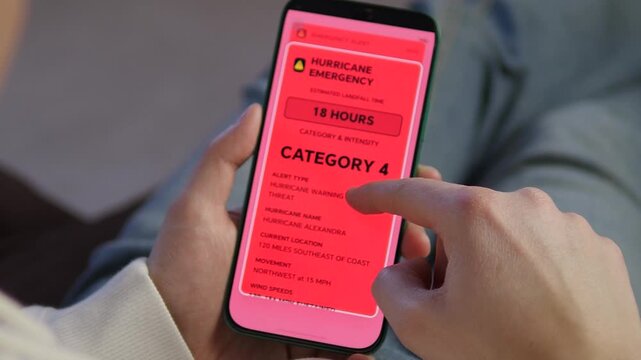 Close-up of hands holding a smartphone with a red Category 4 Hurricane Alexandra emergency alert. Urgent natural disaster notification on mobile screen. Climate change and extreme weather preparedness