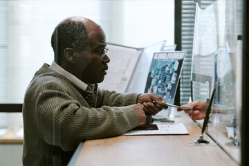 Senior Black man interacting with bank teller, handing over cash and identification card through glass partition at branch counter, financial documents and promotional materials visible in background