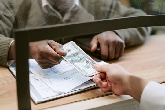 Middle aged Black man handing one hundred dollar bill to woman across bank counter, both hands visible over documents and forms