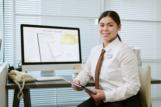 Portrait of young adult woman smiling while sitting at desk in bank branch holding digital tablet, computer monitor with financial information visible in background