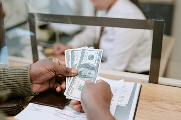 Black man holding US dollar bills at bank branch counter interacting with female bank employee behind glass partition, paperwork and pen on wooden desk in foreground © AnnaStills