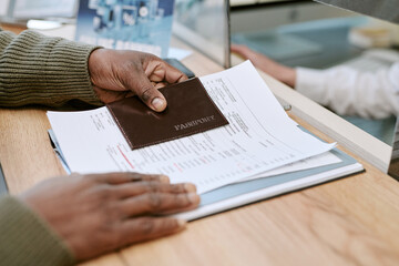 Black man presenting passport and documents at bank branch counter, interacting with employee behind glass partition, close up of hands and paperwork during transaction process