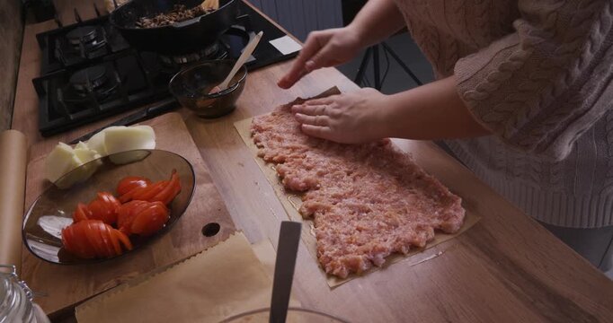 High angle view of a woman's hands carefully flattening minced meat on parchment paper while preparing a homemade meal in a cozy kitchen, with fresh ingredients like tomatoes and cheese nearby.