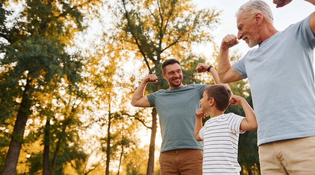 Happy family flexing biceps in sunlit park. Father, son and grandfather connectioneering between three generations of men. Father's Day, copy space
