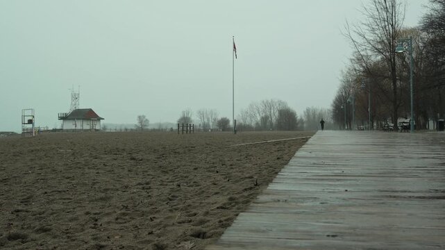 Solitary runner on wet wooden boardwalk in public park, bare trees, flagpole and pavilion visible, overcast rainy morning, Kew Beach, Toronto, Canada.