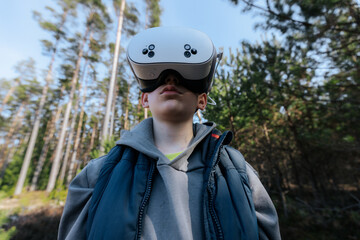 Young boy wearing virtual reality headset stands in a forest, surrounded by tall trees and natural greenery under a clear blue sky © Emvats