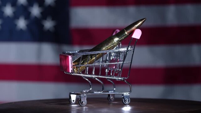 Close up of a bullet placed in a miniature shopping cart against USA flag background symbolizing gun trade consumerism and political debate concept, rotates