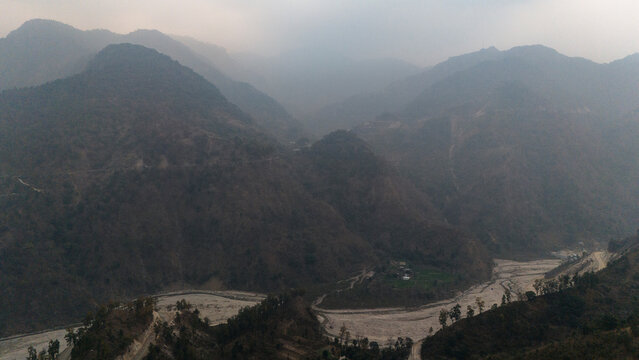 Aerial view of Dwara village near Dehradun, Uttarakhand, with a dirt path along a mountain ridge, misty hills, terrace farms, sal and pine forests, and the Song River below on a winter evening.