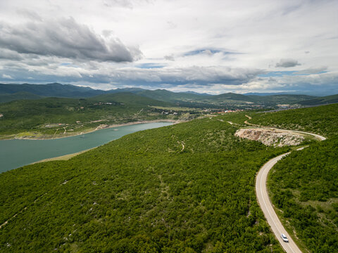 Drone photo of turquoise Bileća Lake, green hills, forest road with car, Henkel site, Torič village, and distant mountains under a cloudy sky.