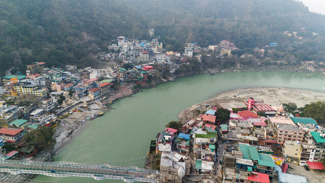 Aerial view of Lakshman Jhula in Rishikesh, Uttarakhand, with the Ganga River flowing below, and temples, houses, and buildings along the riverbank against misty mountains on a winter morning.