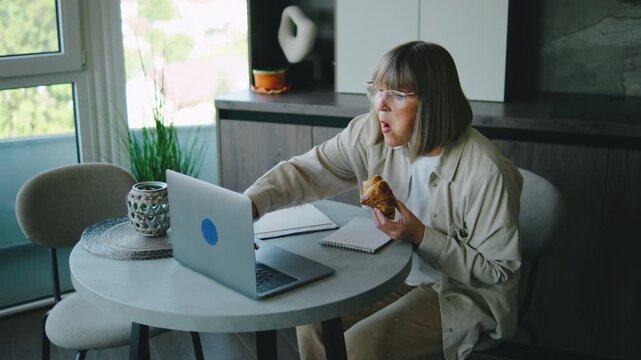 Elderly woman sits at a table with a laptop, writing notes and eating a pastry in a bright room with plants and a city view