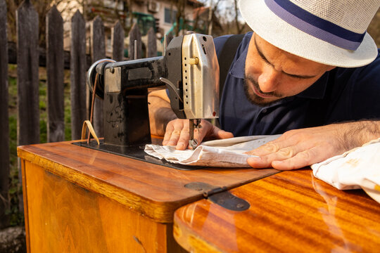 Man carefully sewing fabric on vintage machine outdoors, leaning over wooden table with focused expression, capturing dedication and precision in traditional tailoring craft.