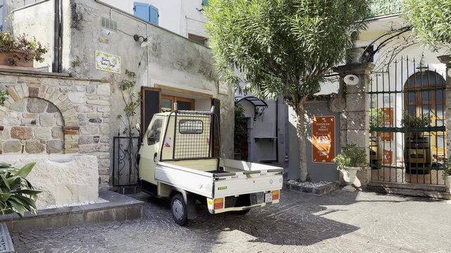 Yellow Piaggio Ape Three Wheeler Parked in Charming Courtyard with Stone Wall Iron Gate Oleander Trees and Local Shop in Limone sul Garda Italy 4K HDR